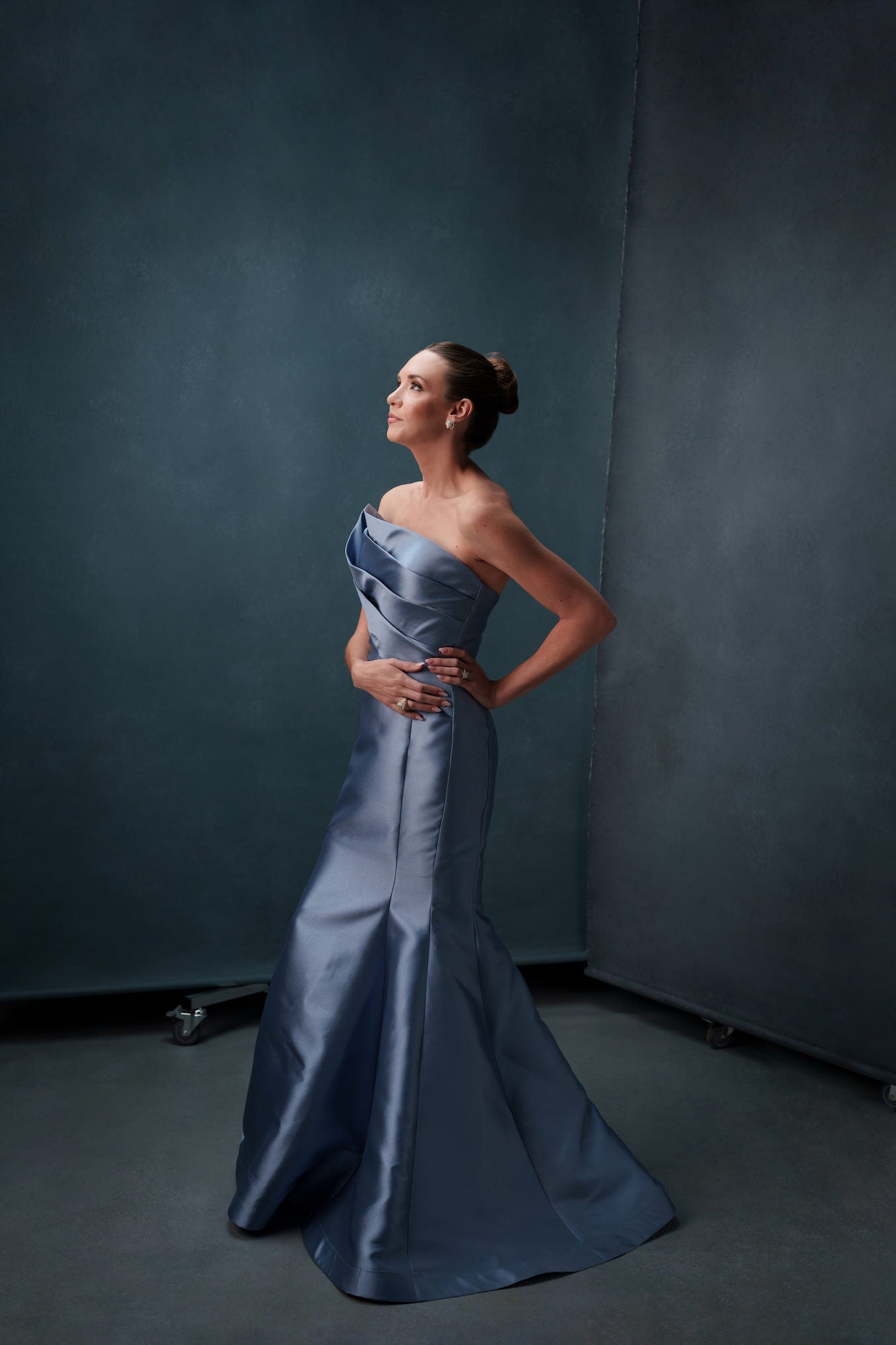 Woman in a strapless slate‑blue evening gown posing in profile with hands on hips against a textured studio backdrop. Woman in a strapless slate‑blue evening gown posing in profile with hands on hips against a textured studio backdrop.