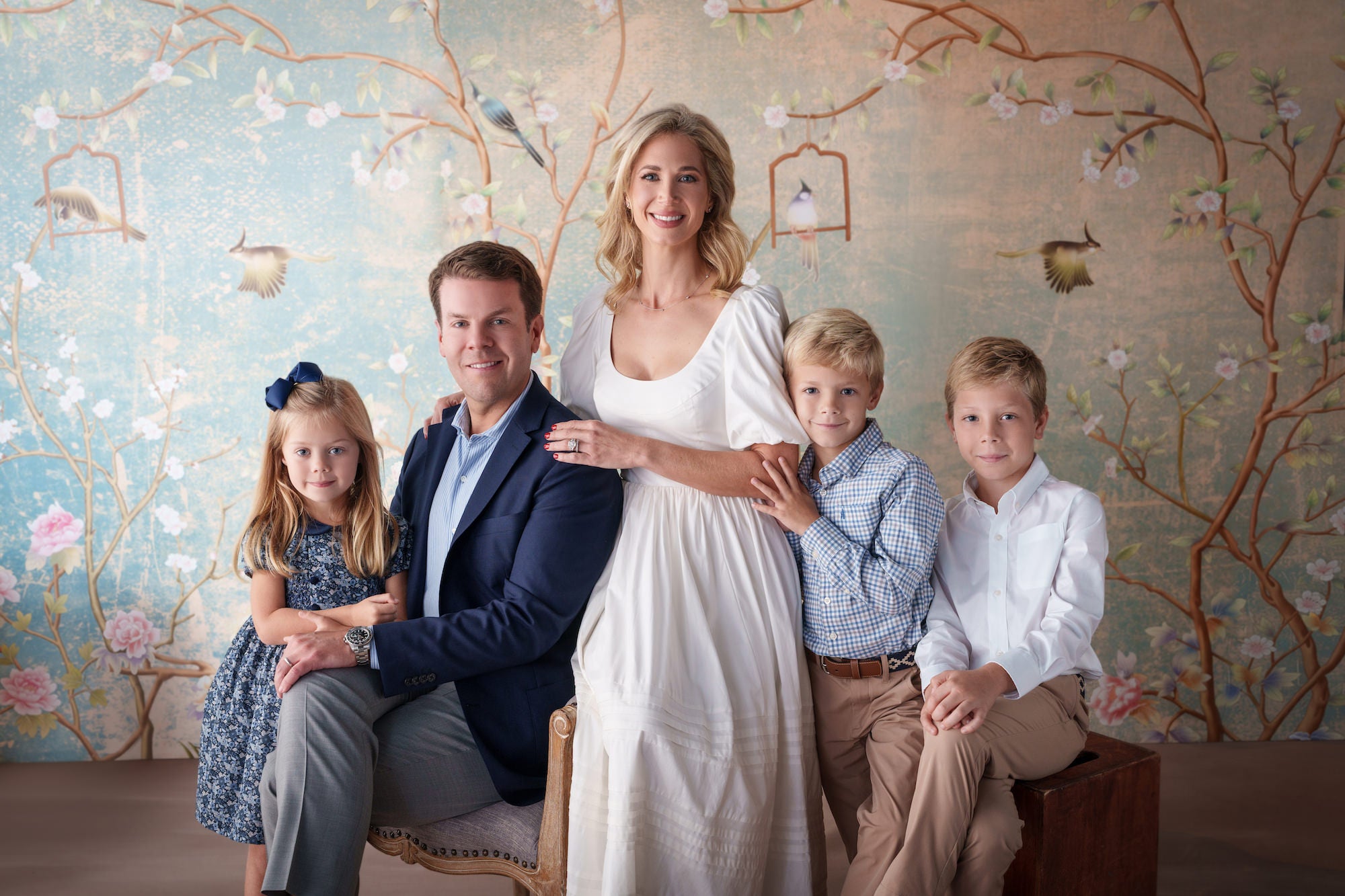 Studio family portrait of five in soft blues and whites, posing in front of a pastel bird‑and‑floral mural backdrop. Studio family portrait of five in soft blues and whites, posing in front of a pastel bird‑and‑floral mural backdrop.