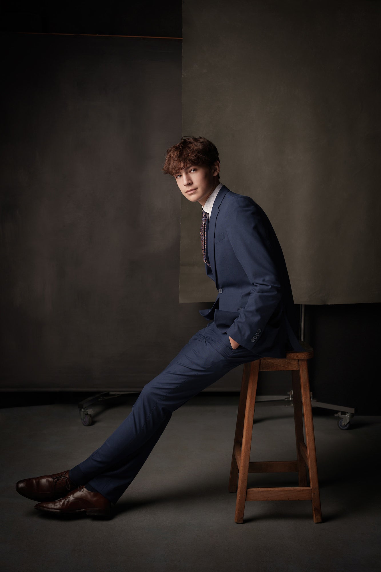 Young man in a blue suit seated on a wooden stool in a studio, legs extended and hands in pockets, moody backdrop. Young man in a blue suit seated on a wooden stool in a studio, legs extended and hands in pockets, moody backdrop.
