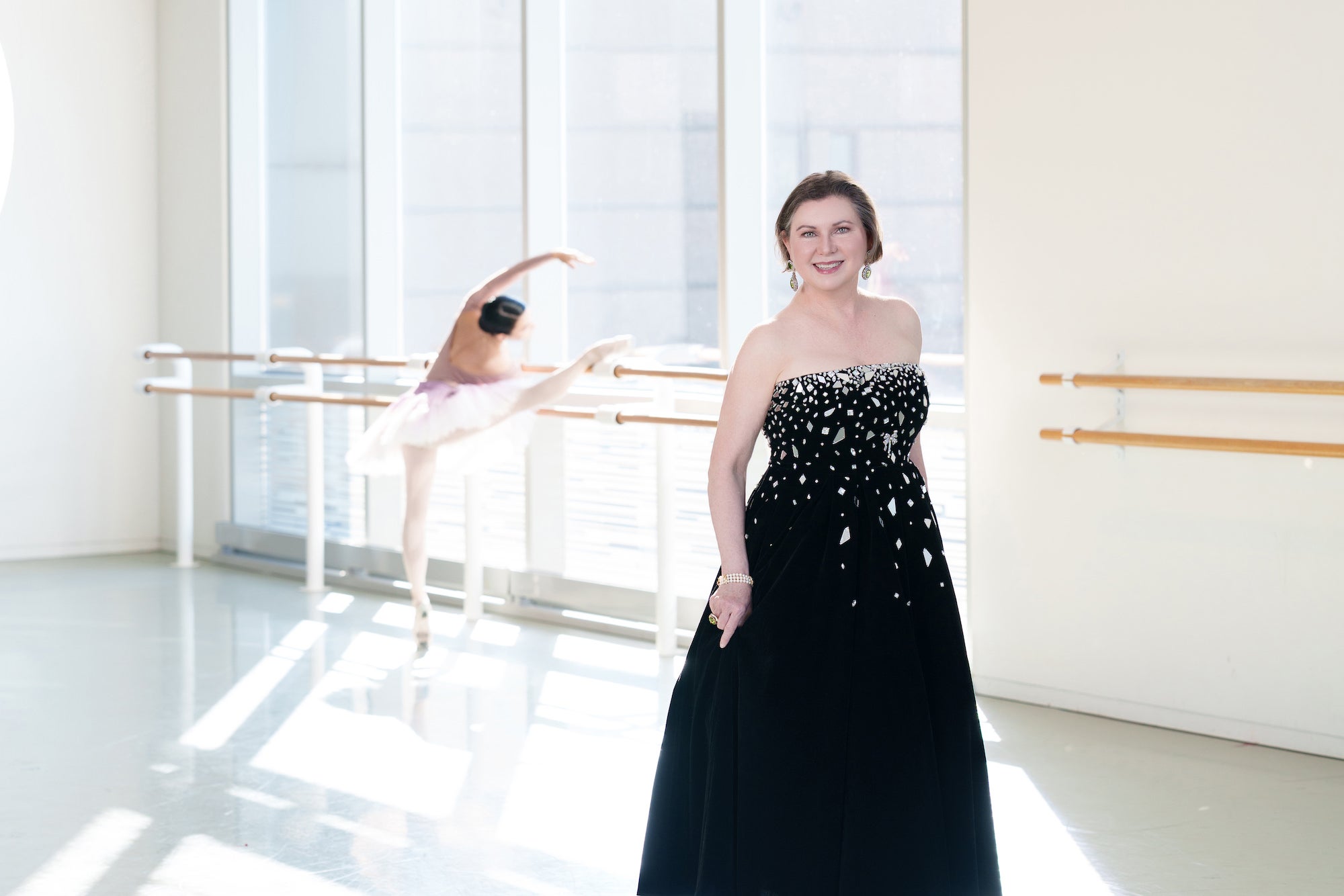 Woman in a black strapless gown in a bright ballet studio; dancer blurred in the background at the barre. Woman in a black strapless gown in a bright ballet studio; dancer blurred in the background at the barre.