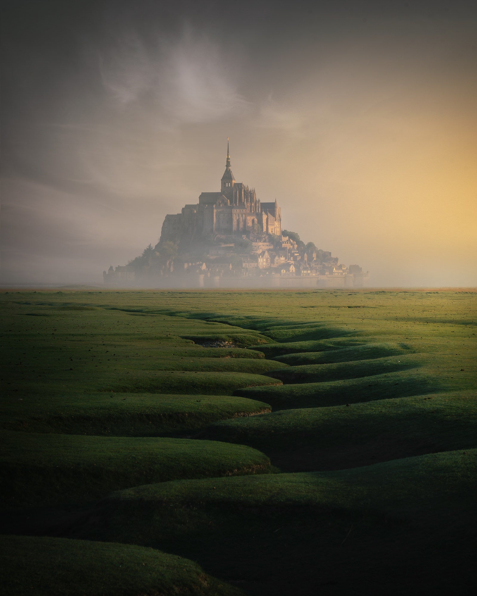 Mont-Saint-Michel rises from coastal fog at sunrise with sculpted tidal meadows in the foreground.