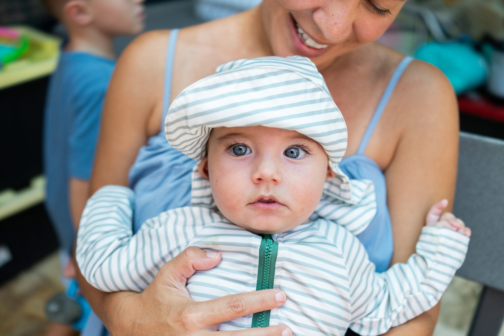 Close-up of a baby in a striped sun outfit with blue eyes, held by a smiling mother.