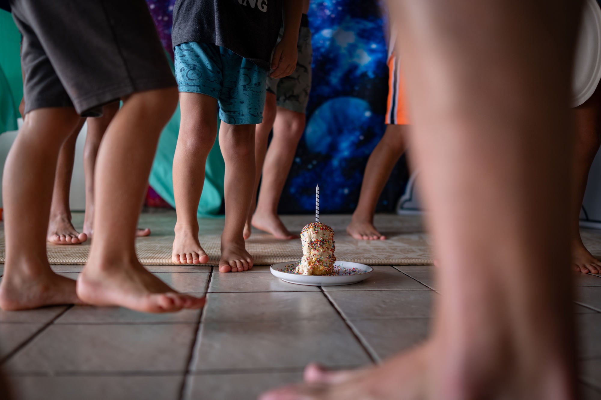 Children gather barefoot around a sprinkle-covered cupcake with a candle, space backdrop behind.