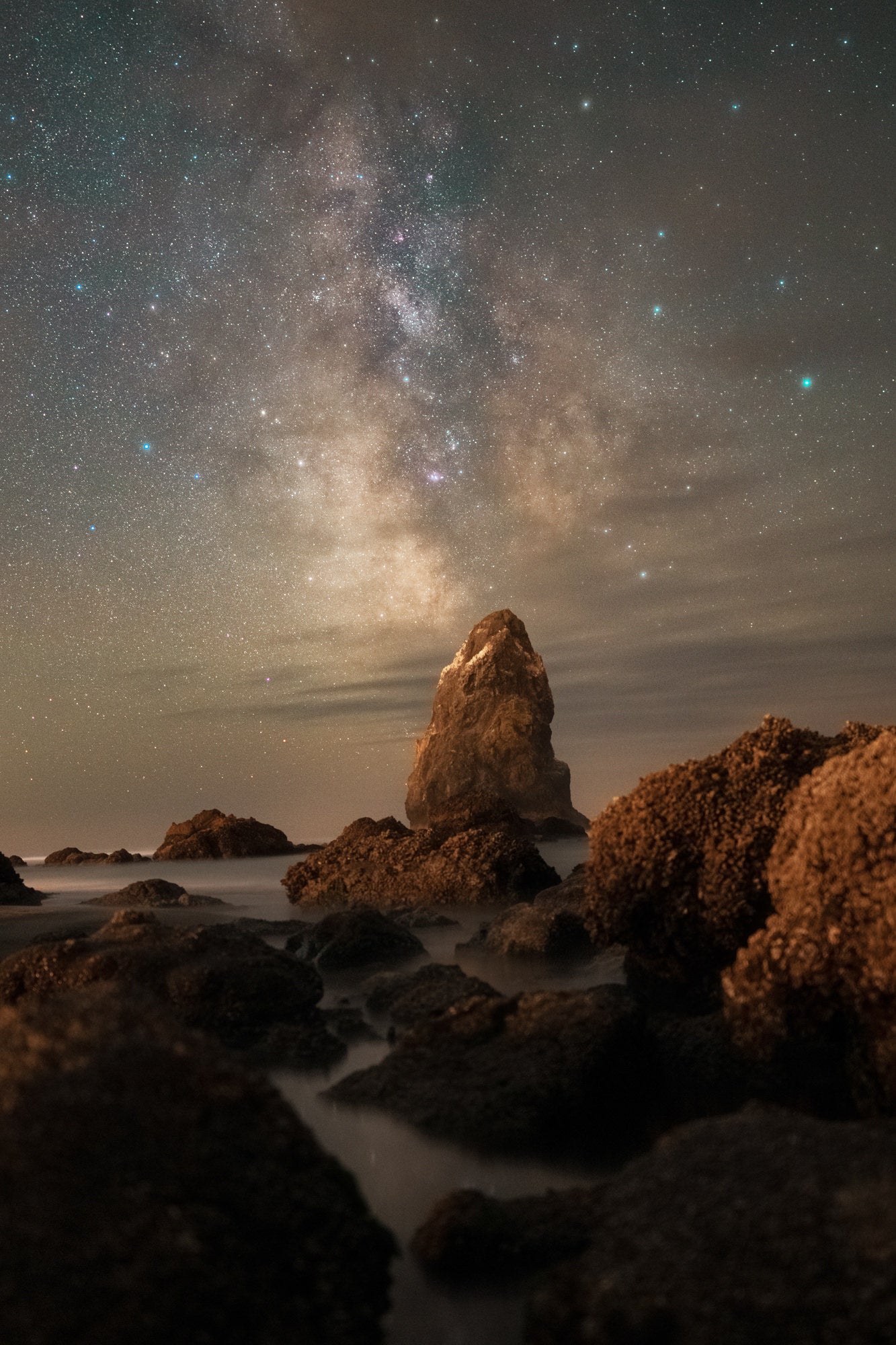 The Milky Way arching over a sea stack and tide-pool rocks at Cannon Beach, with surf blurred by long exposure.