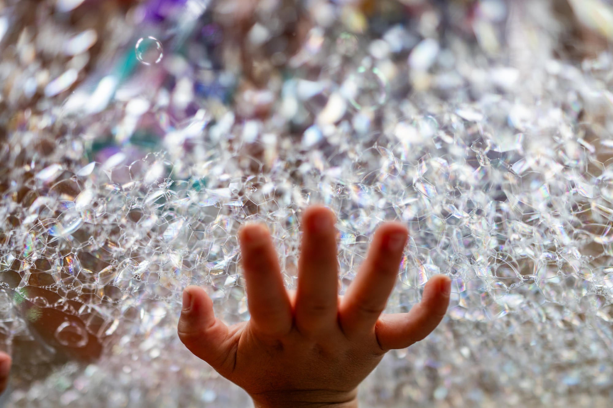 Small hand reaches into a sparkling mass of soap bubbles with colorful bokeh highlights.