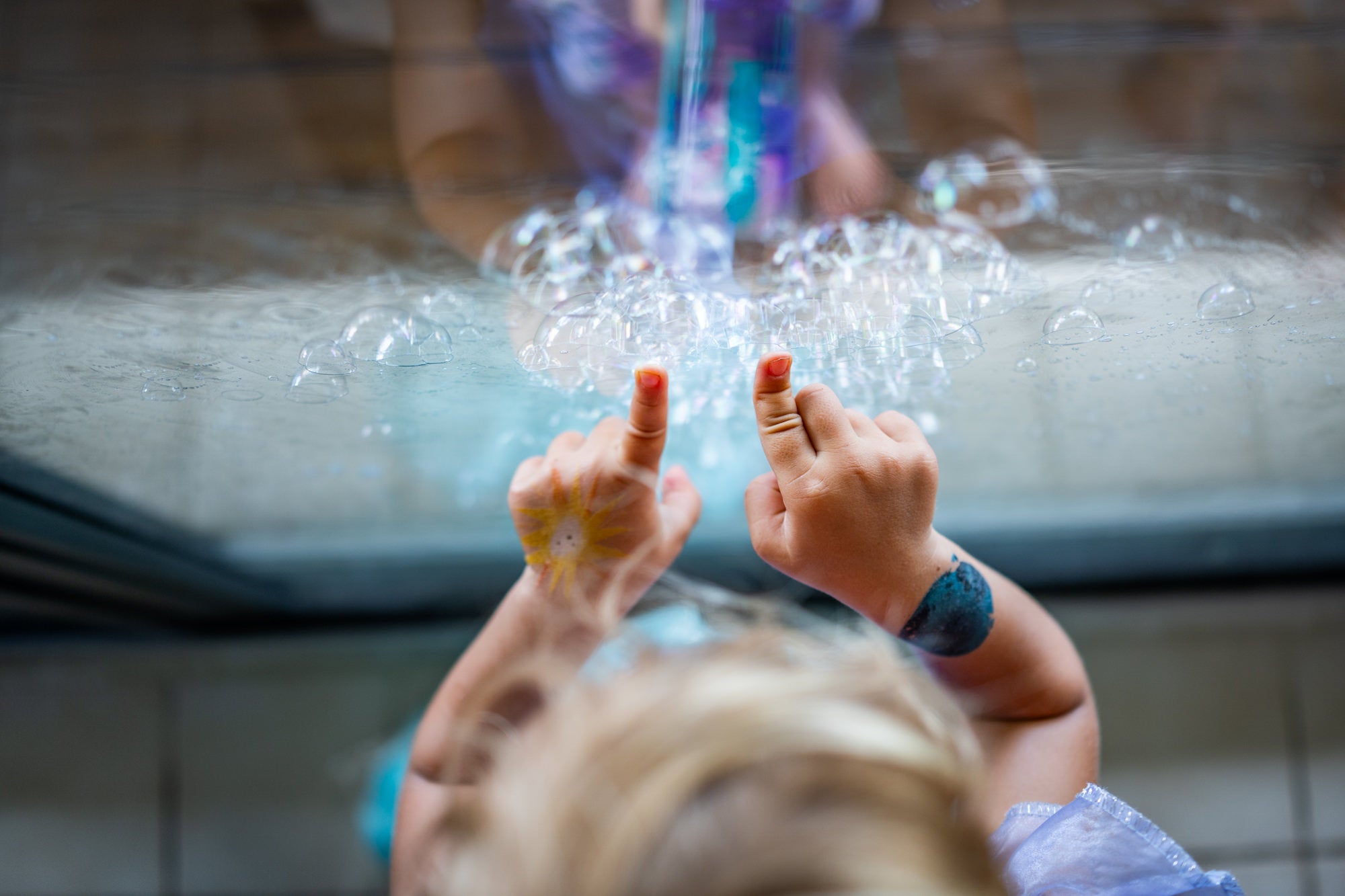 Child’s hands poke bubbles on a glass surface with small sun and planet drawings on skin.