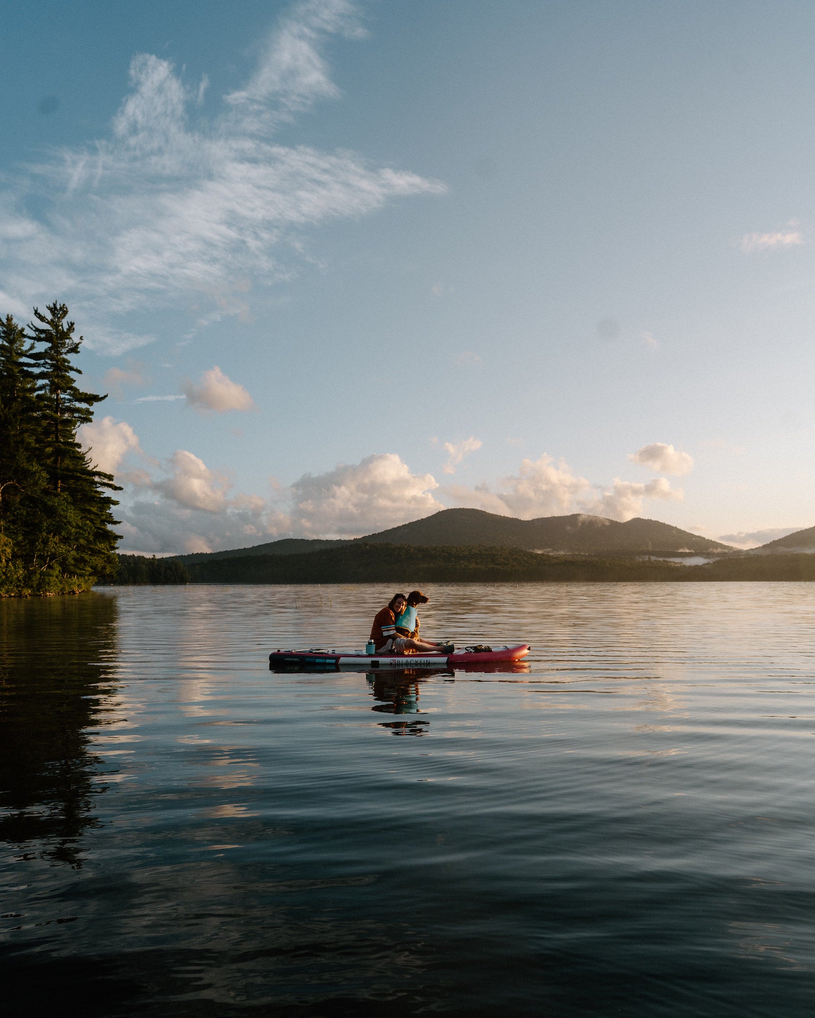 Couple sitting close together on a paddleboard in the middle of a calm lake with pines and mountains at sunset. Photo by Rachael Austin. 24mm f/1.4 G Master, f/5, 1/1000-sec. ISO 200.