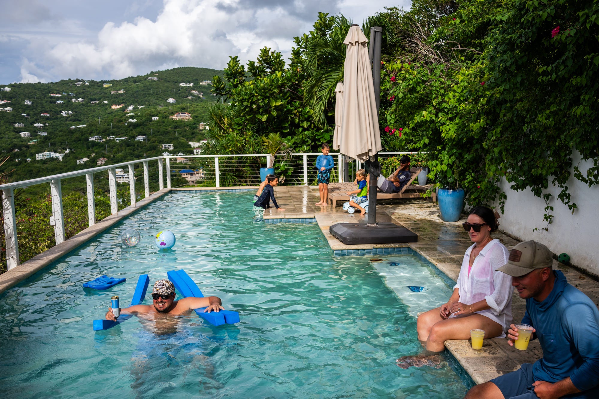 Family and kids enjoying a hillside pool with floats, drinks, and lush tropical views.