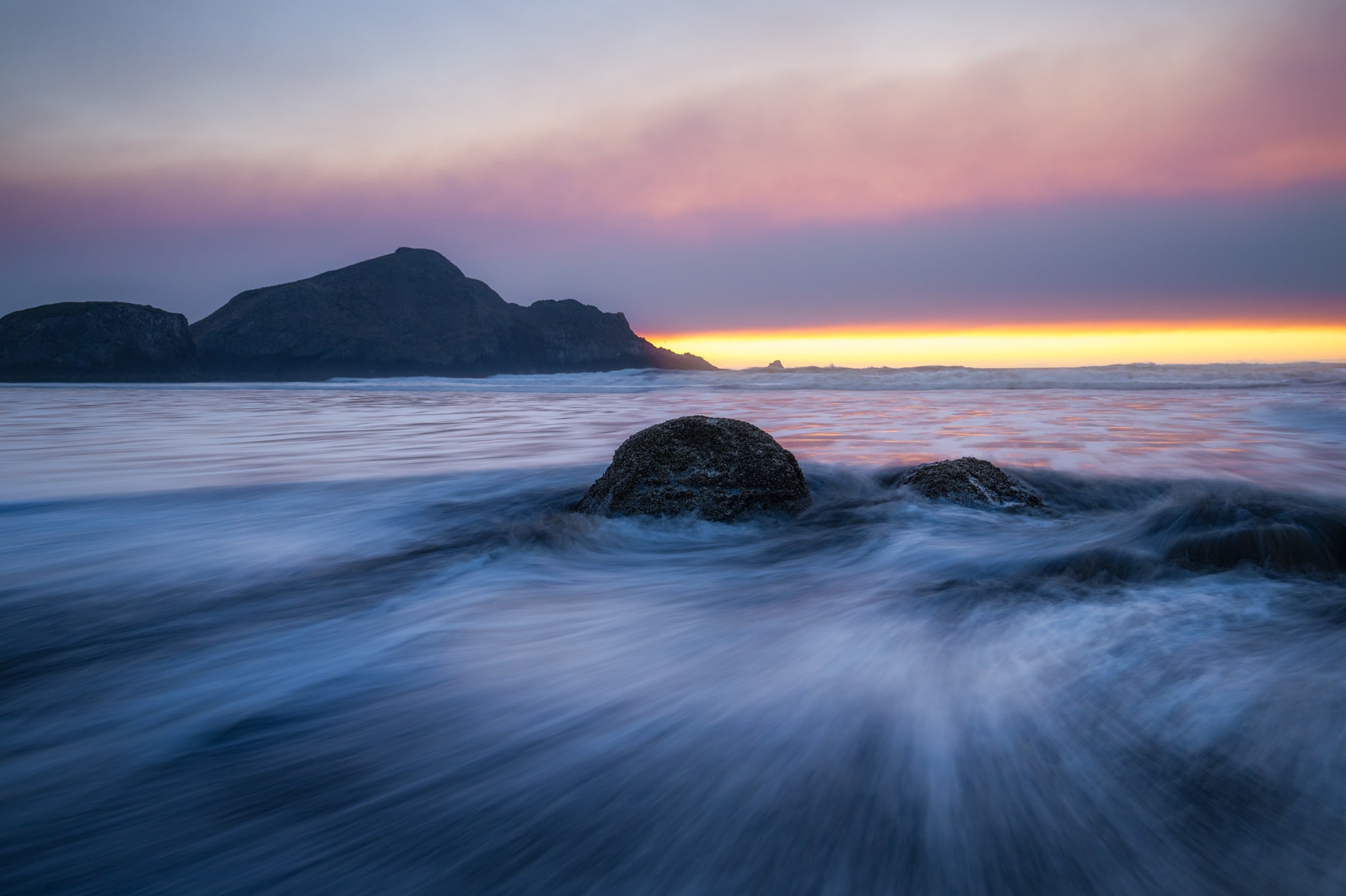 Long exposure of surf streaming past rocks at Gold Beach, with a dark headland under a pink and gold sunset sky.