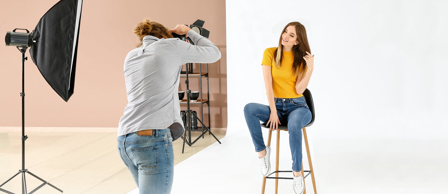 A photographer takes a portrait of a woman wearing a yellow sweater and blue jeans. She is sitting on a stool in front of a white background. There is a strobe with a softbox to the left of the photographer.