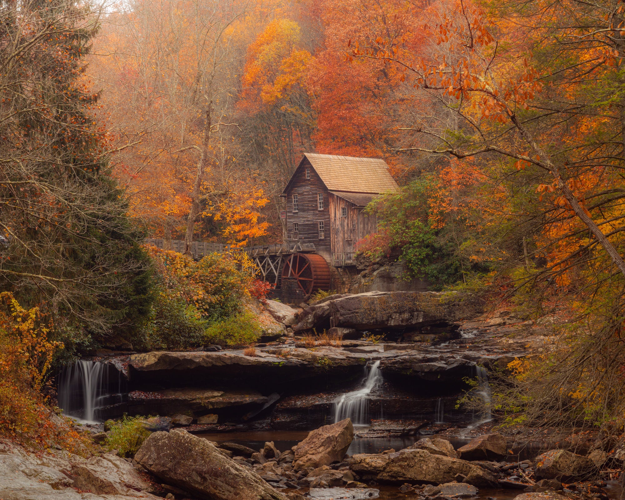 Wooden grist mill above a silky long-exposure creek and rock ledges, framed by orange autumn forest in fog. Photo by David Shockey.