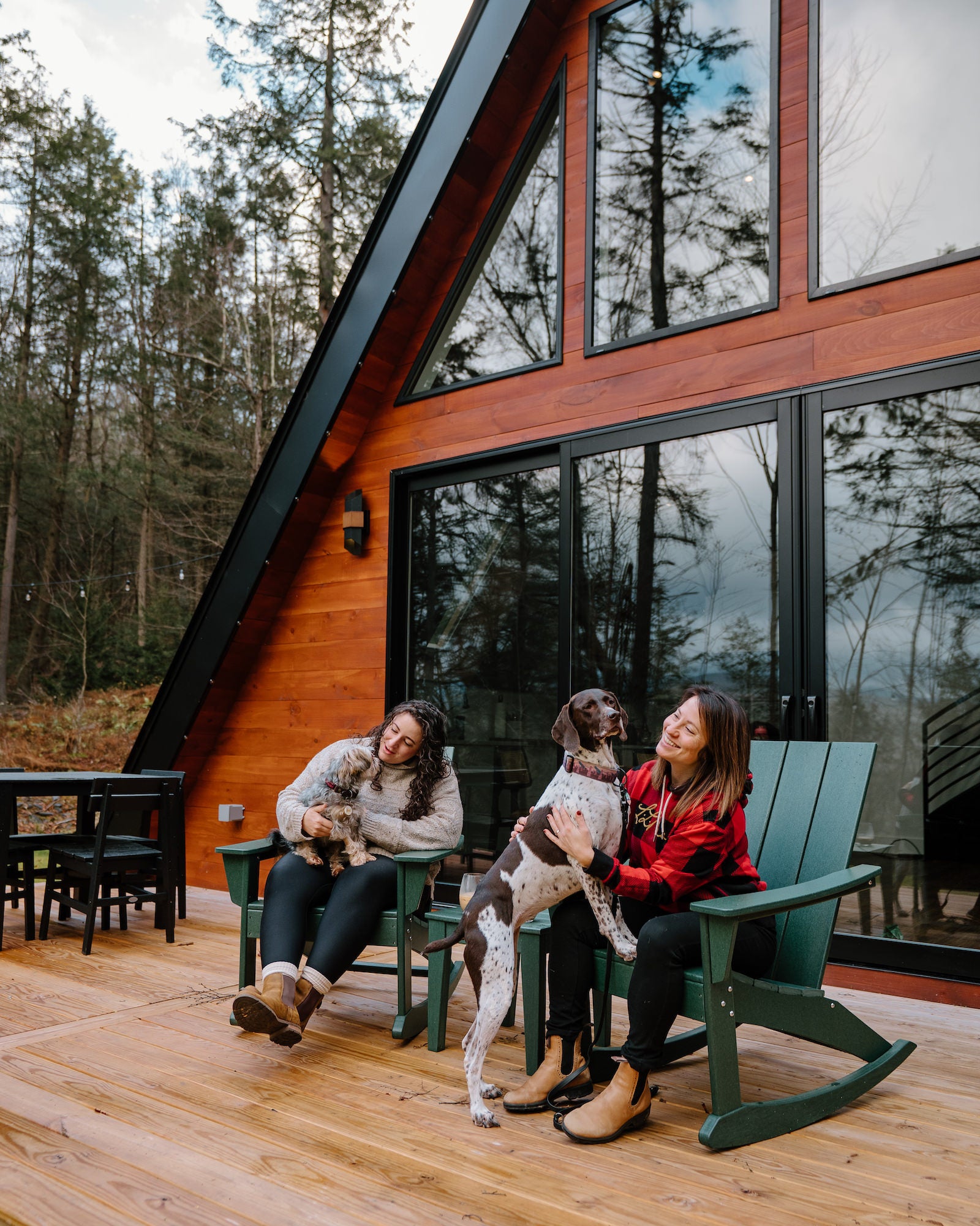 Two women sit in green Adirondack chairs with their dogs on the deck of an A-frame cabin in the woods. Photo by Rachael Austin. 16-35mm f/2.8 G Master, f/2.8. 1/400-sec. ISO 400.