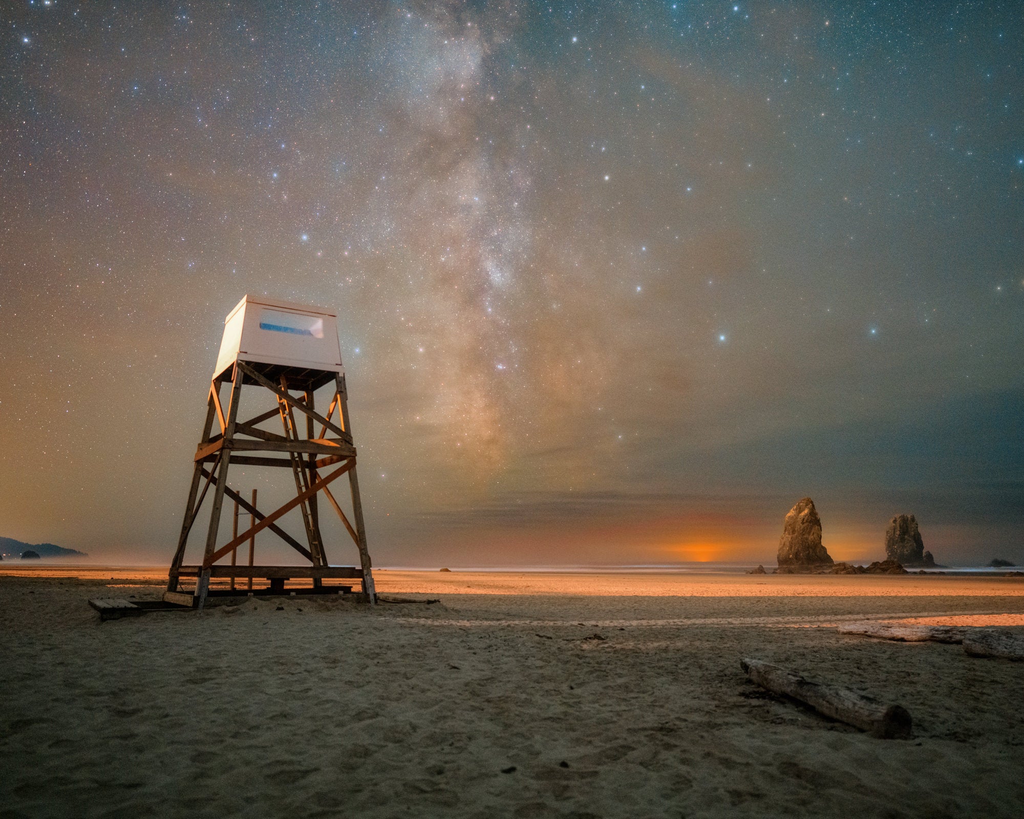 Milky Way arcing over a wooden lifeguard tower on a sandy beach with sea stacks and glowing horizon. Photo by David Shockey.