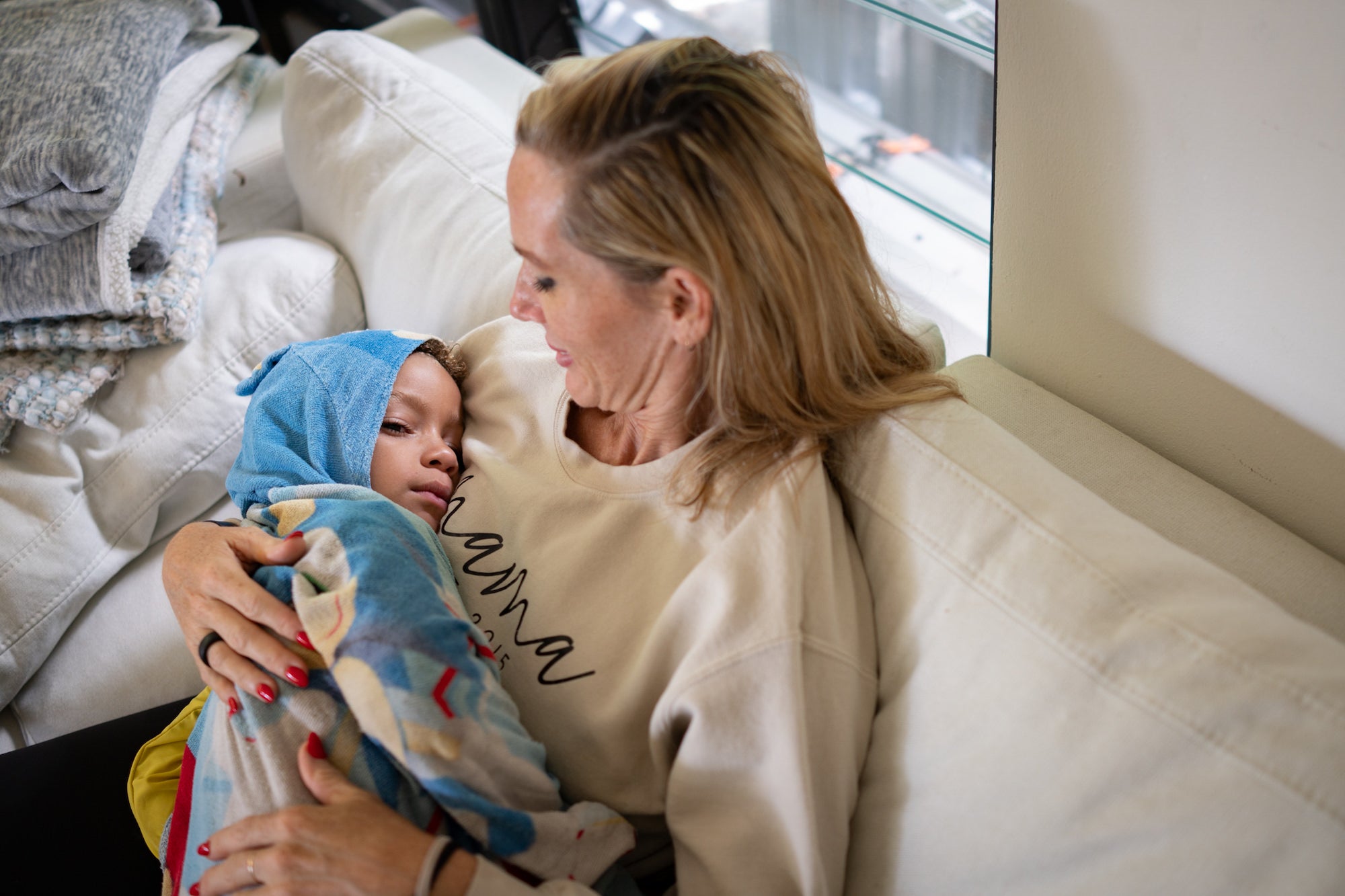 Mother holding a blanket-wrapped child on a white couch, sharing a quiet, comforting moment.