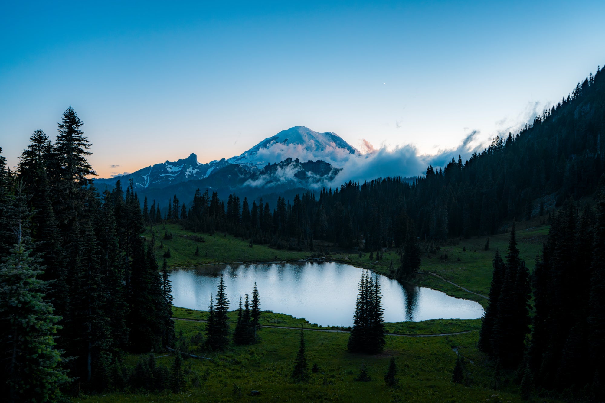 Alpine lake ringed by evergreens and meadow at dusk, with a snow-capped, cloud-wrapped peak in Mount Rainier National Park. Photo by Tomer Negrin. Sony Alpha 7R V. 16-35mm f/2.8 G Master II. f/4.5. 1/3-sec. ISO 800.