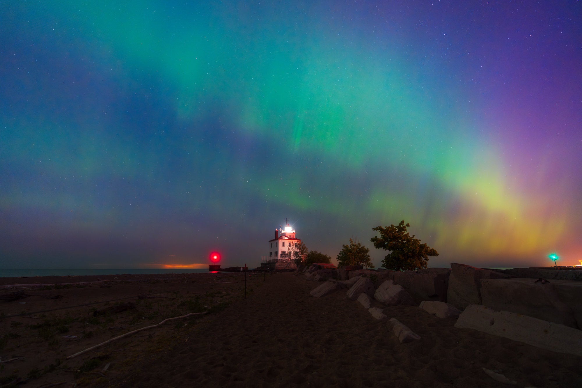 Green and purple northern lights fill the night sky above a lit lighthouse on a rocky beach shoreline. Photo by David Shockey.