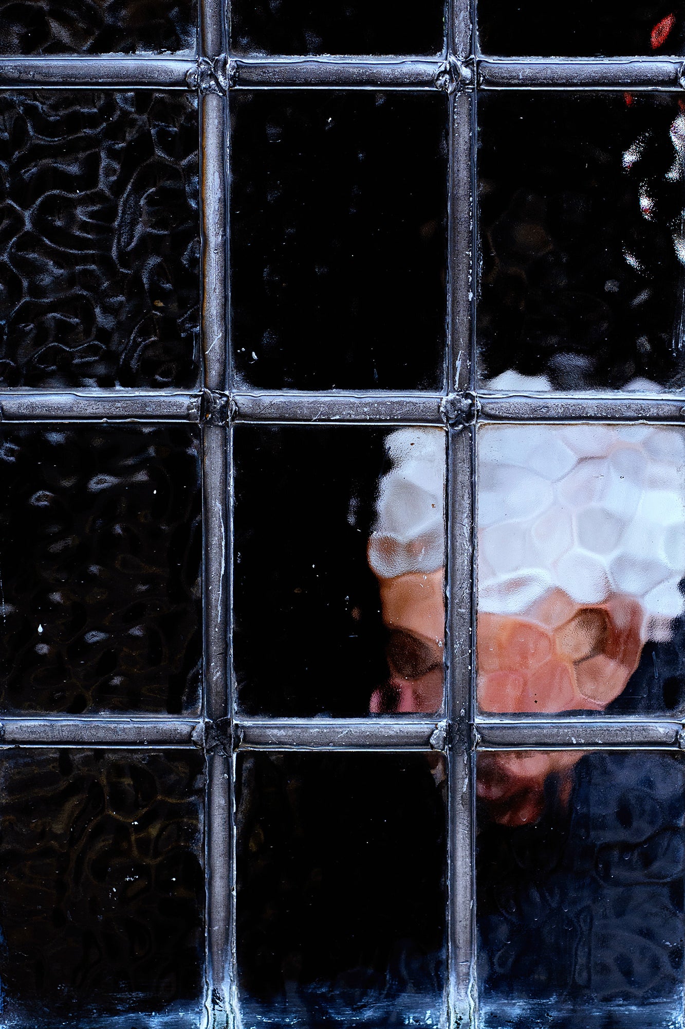 Obscured face framed by textured window panes, dark moody portrait.