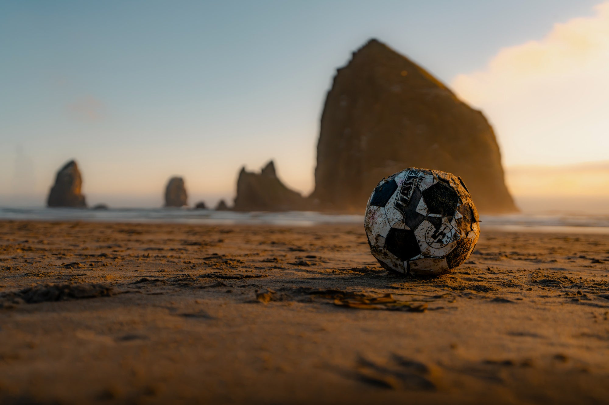 Weathered soccer ball resting on sand at low angle, with sea stacks blurred in the background at sunset. Photo by Tomer Negrin. Alpha 7R V. 16-35mm f/2.8 G Master II. f/2.8. 1/5000-sec. ISO 200.