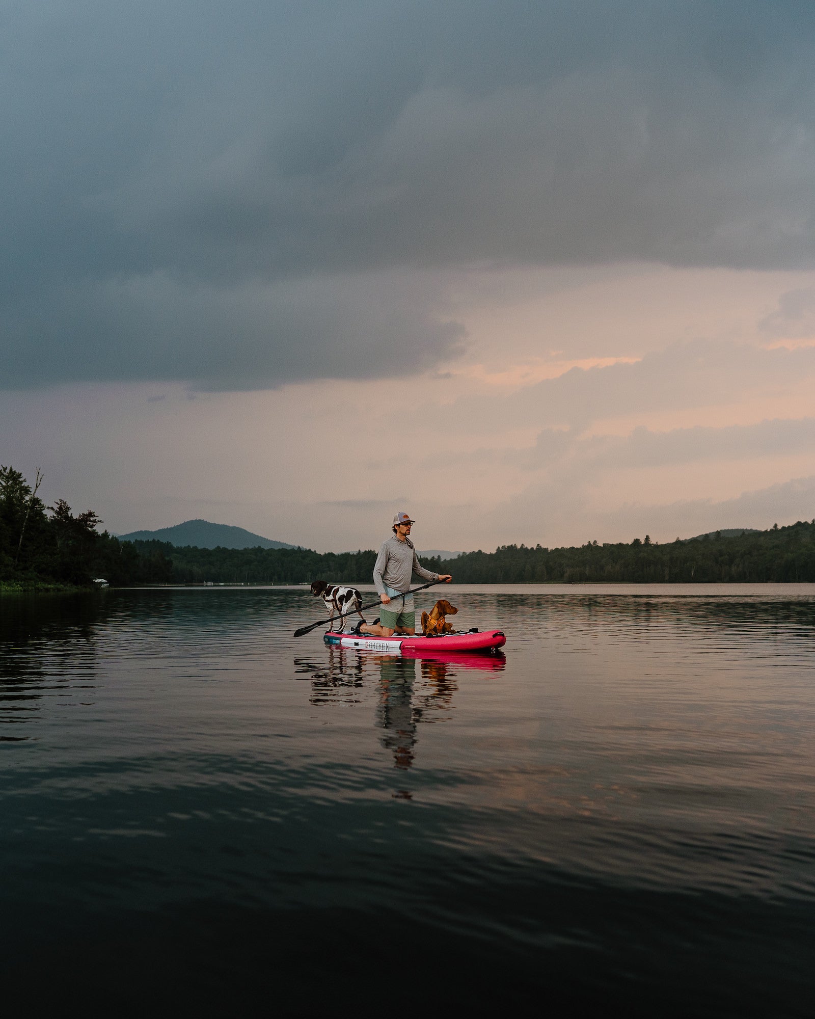 Man paddleboarding with two dogs on a calm mountain lake under stormy clouds at dusk. Photo by Rachael Austin. 24mm f/1.4 G Master, f/2.5, 1/320-sec, ISO 200.