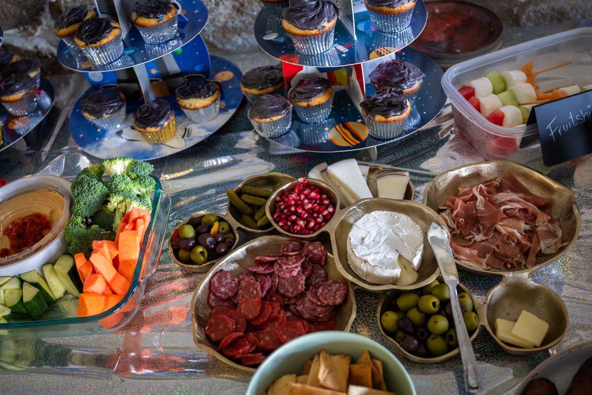Space-themed party table with cupcakes, charcuterie, veggies, hummus, and fruit skewers.