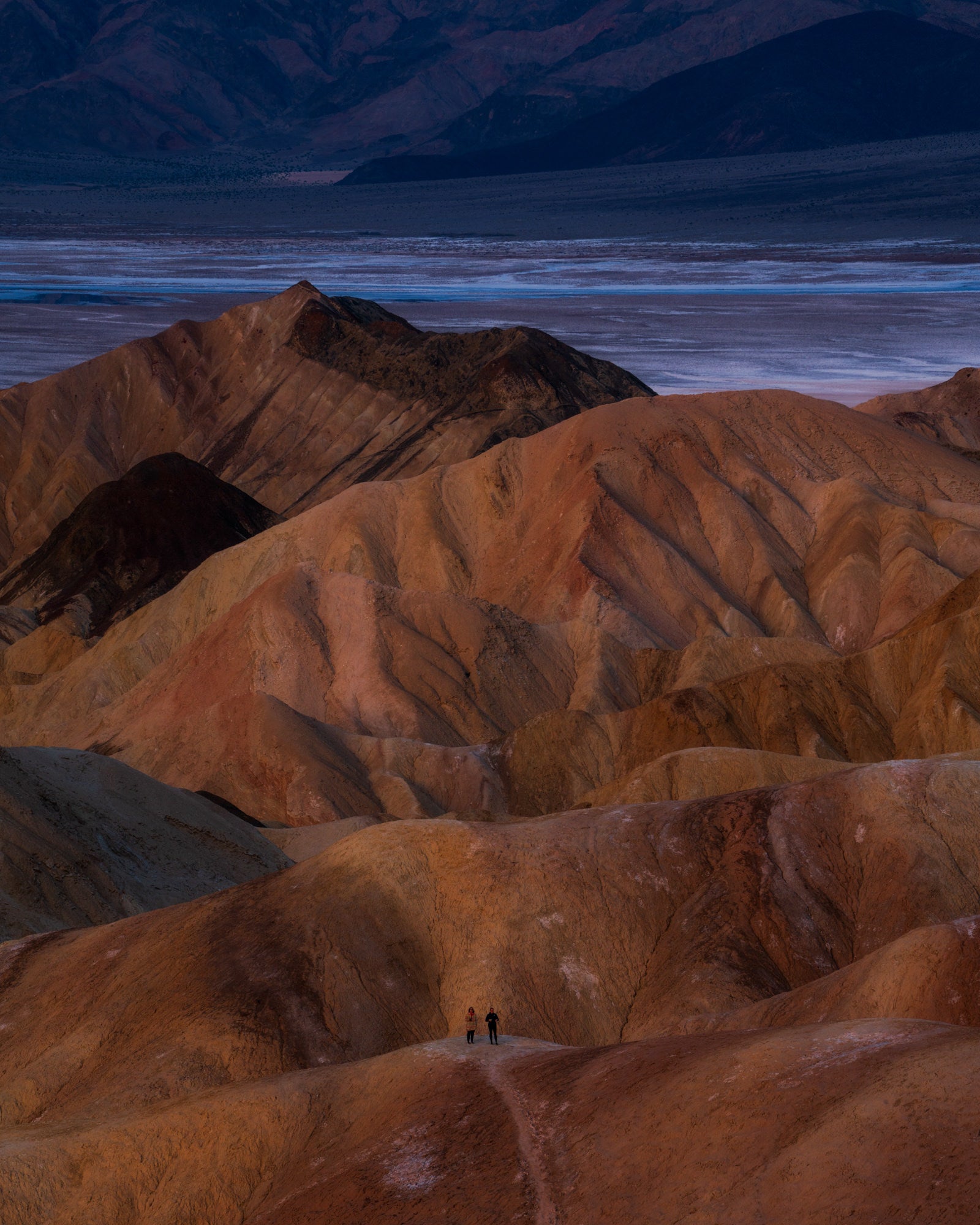 Two tiny figures stand on a ridge amid vast eroded golden badlands with salt flats and dark mountains beyond. Photo by David Shockey.