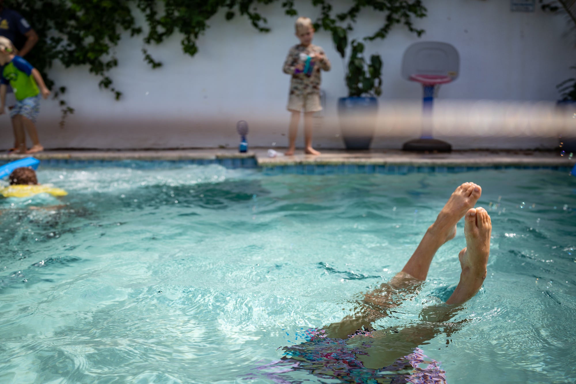 Poolside scene with kids playing as a swimmer’s legs kick above the water’s surface.