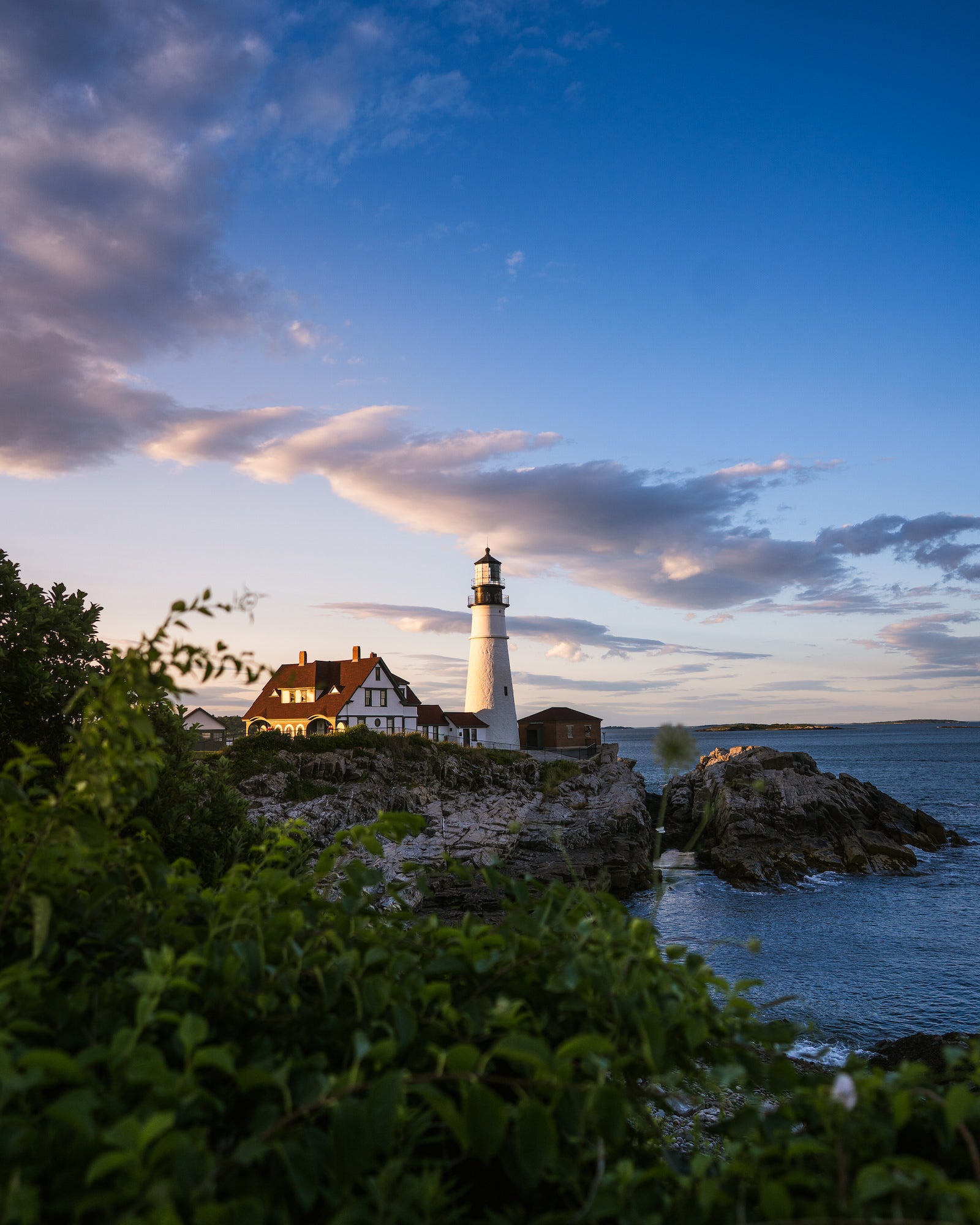 Portland Head Light and keeper's house glowing at sunset above rocky coastal cliffs, framed by green foliage. Photo by Rachael Austin. 24mm f/1.4 G Master, f/2.2, 1/2000-sec, ISO 100.
