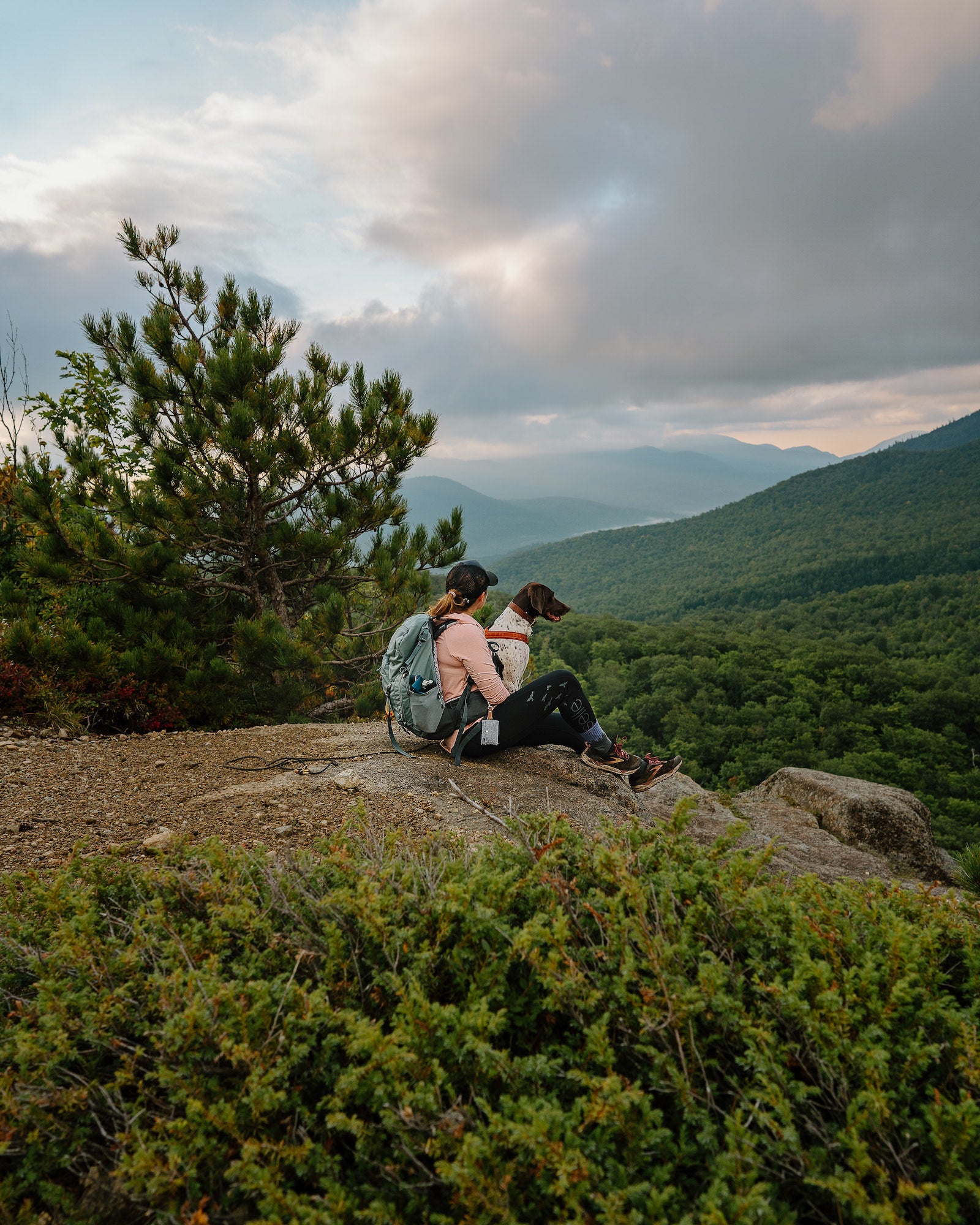 Hiker and her dog resting on a rocky summit ledge overlooking forested mountain ridges at Owl's Head. Photo by Rachael Austin. 24mm f/1.4 G Master, f/4.5, 1/500-sec, ISO 250