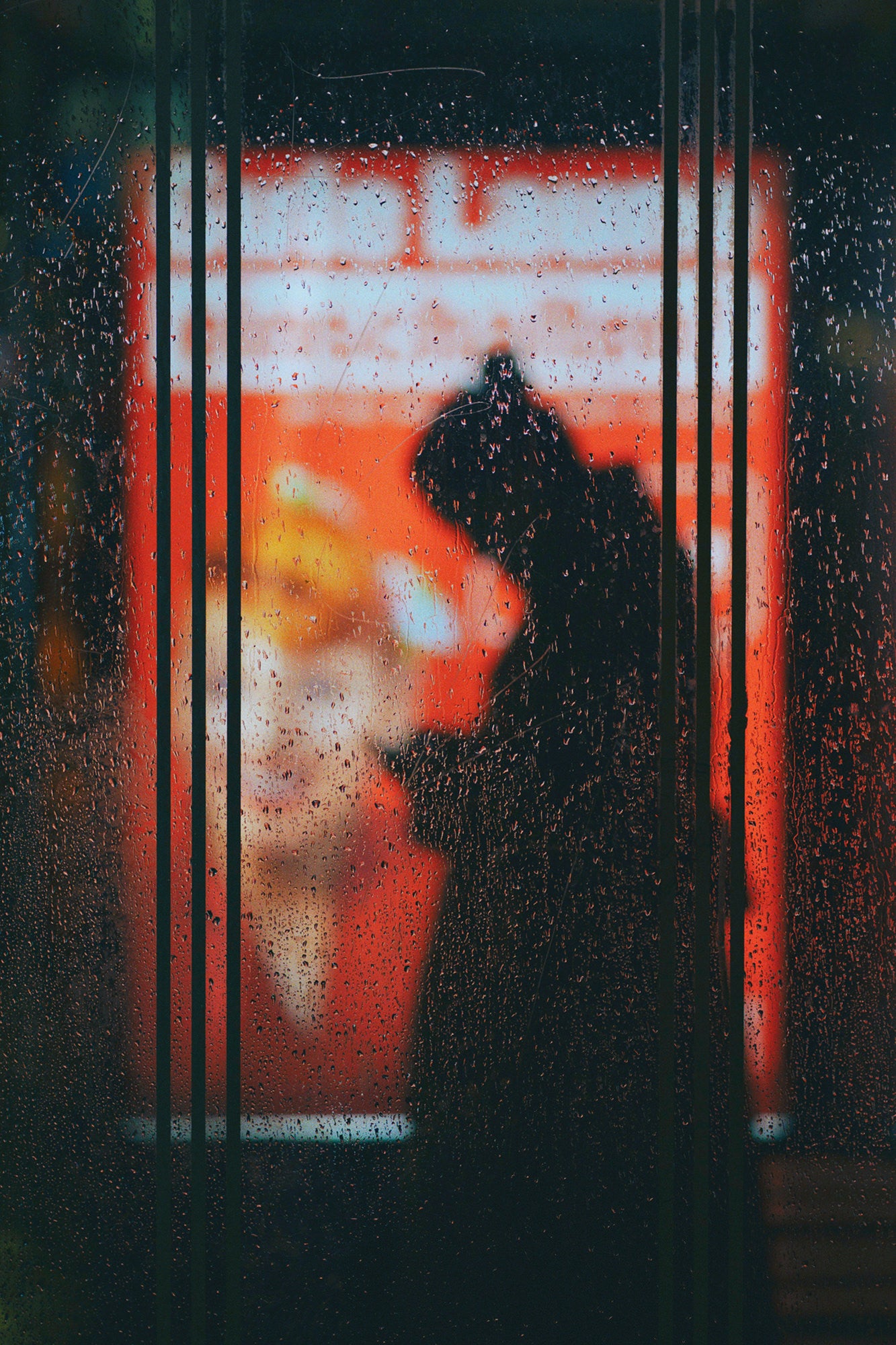 Silhouette at a rainy bus stop seen through wet glass and orange signage.