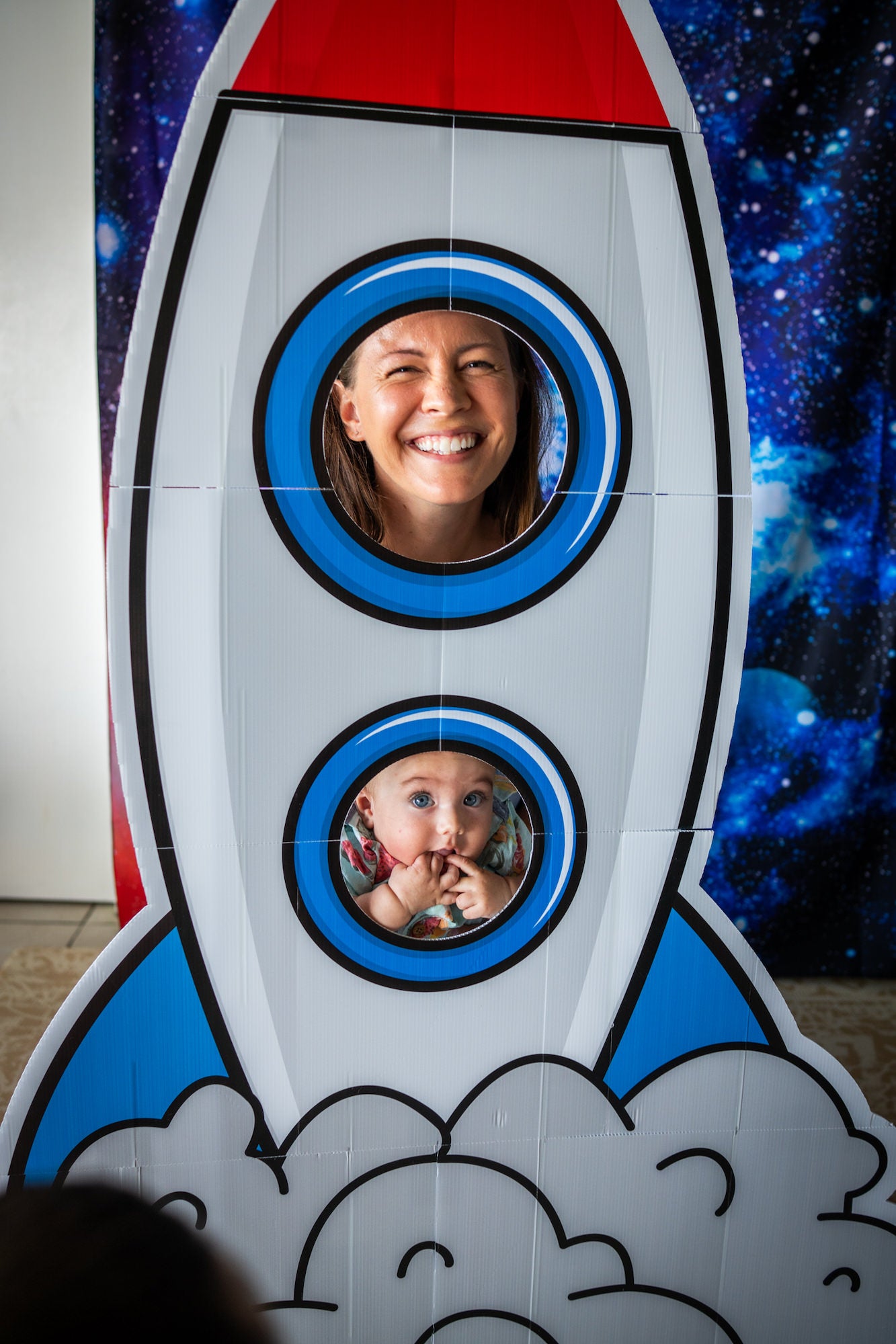Woman and baby smile through rocket photo booth cutouts against a starry galaxy backdrop.
