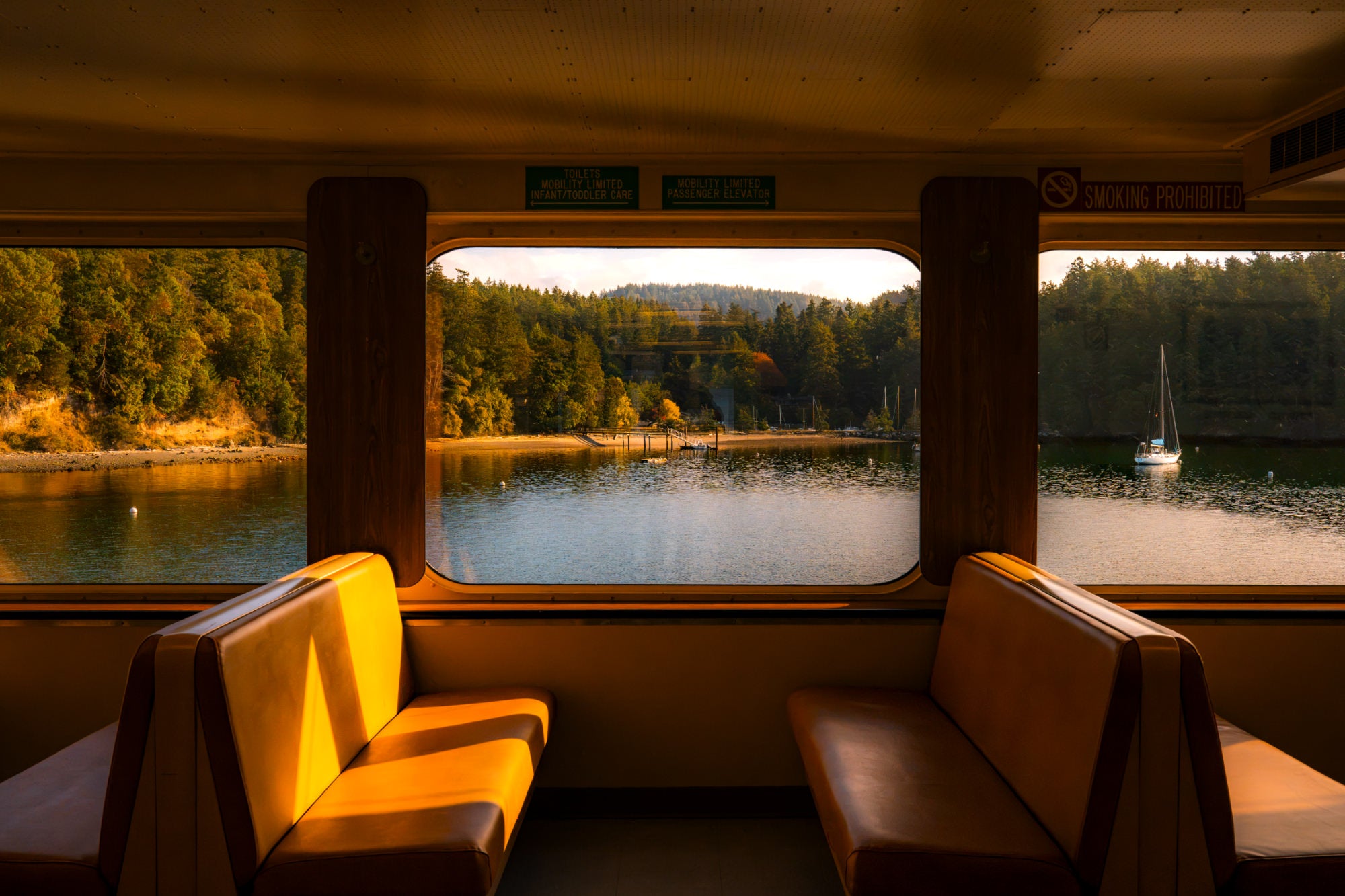 Empty ferry cabin seats in golden light, windows framing a forested shoreline and anchored sailboat.