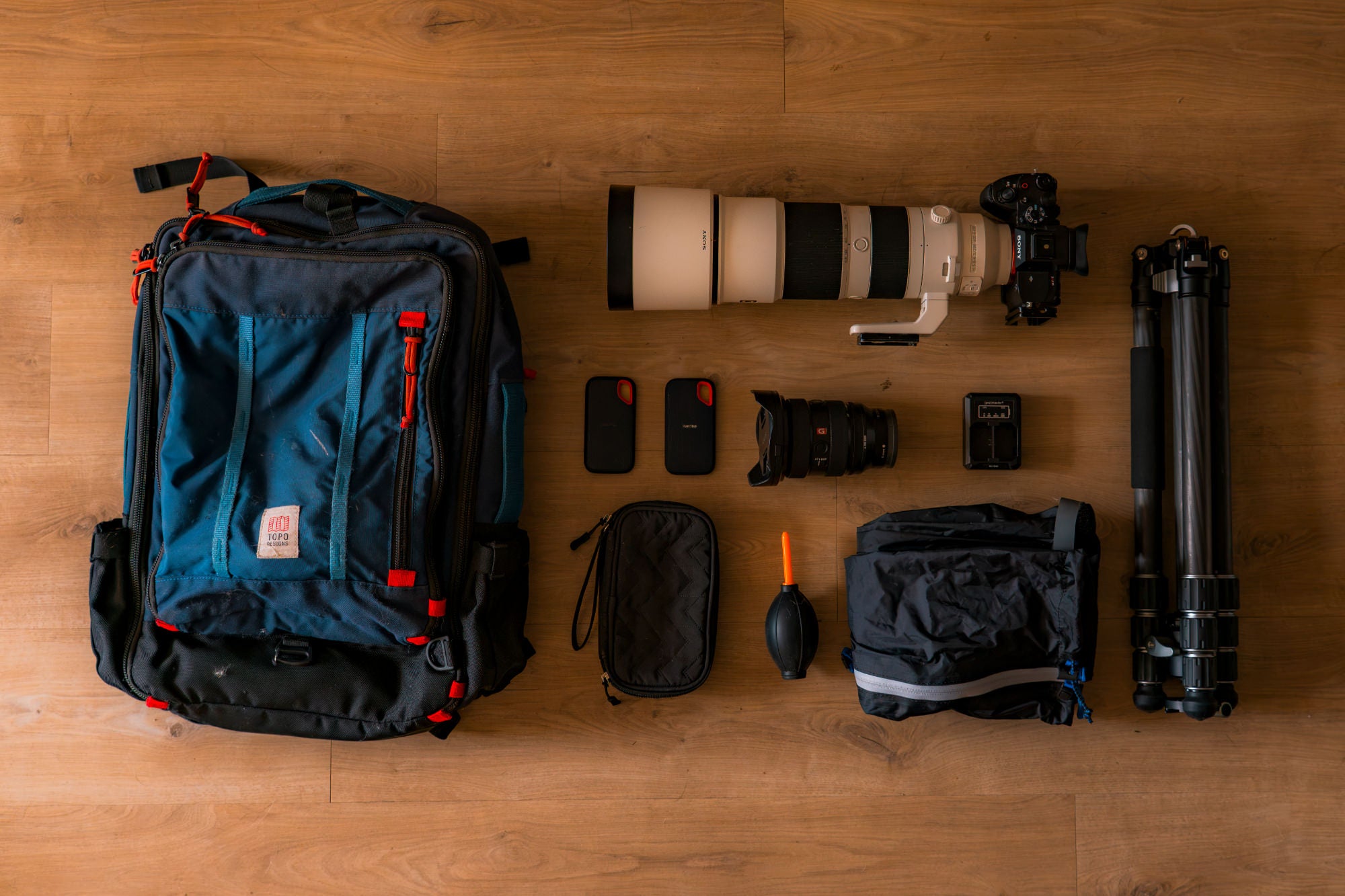 Flat lay of camera gear on a wood floor: backpack, Sony body with white telephoto lens, wide zoom, SSDs, blower, tripod.