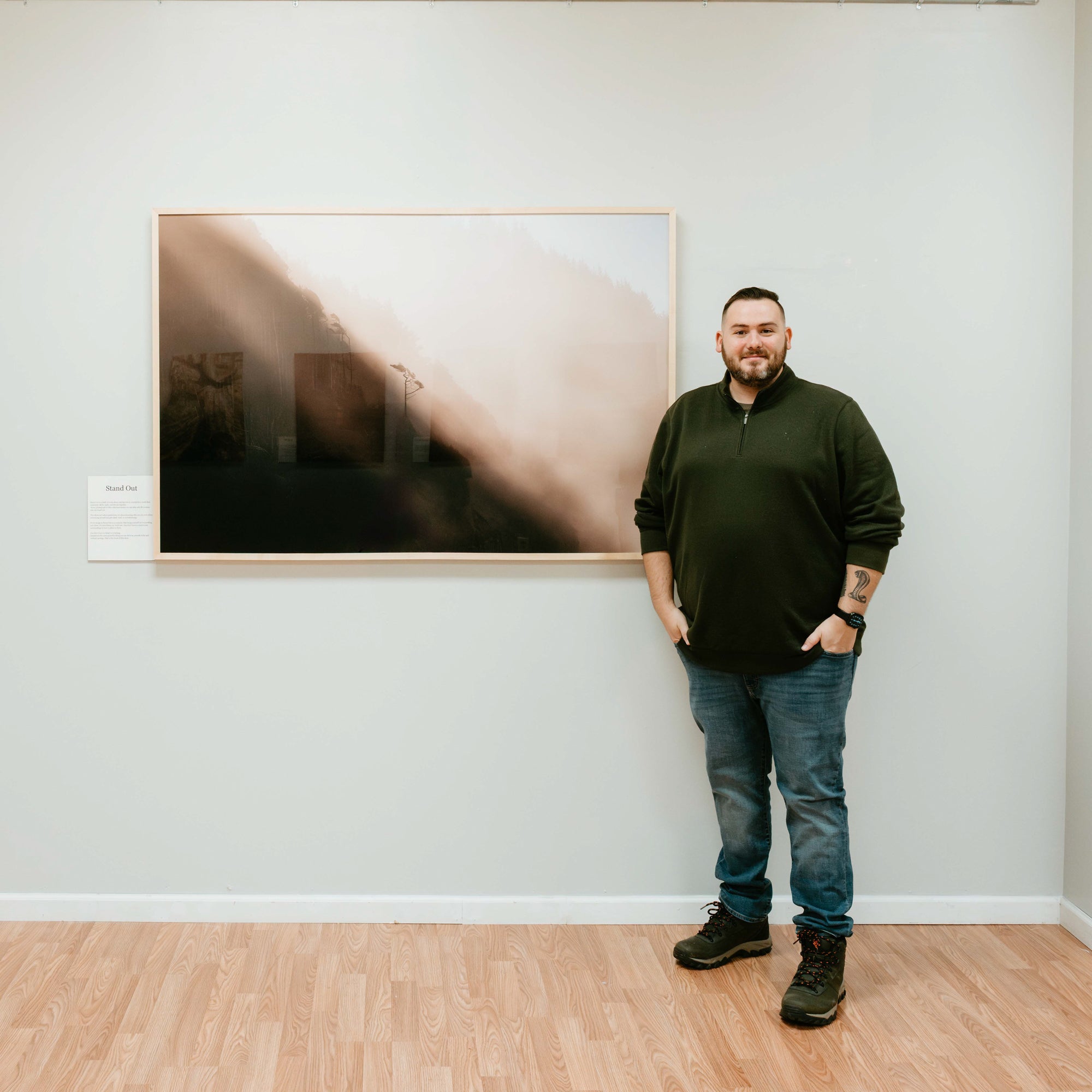 Man in a dark pullover and jeans stands in a gallery beside his large framed misty landscape print titled 