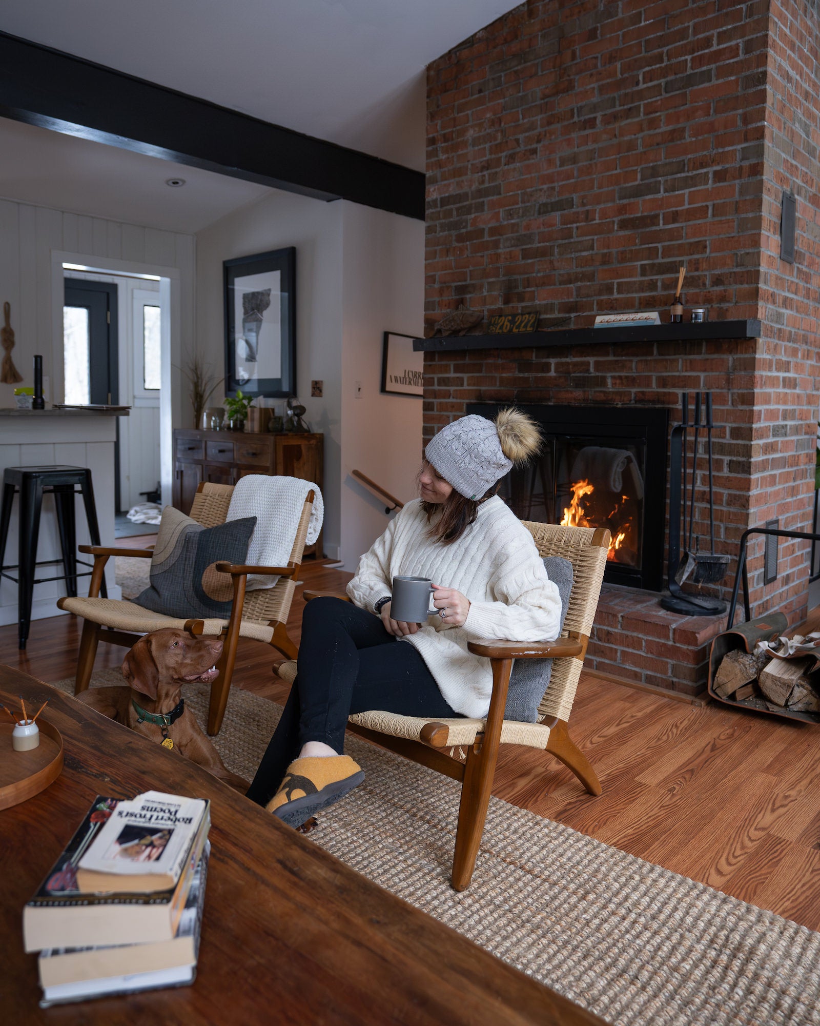 Woman in knit hat holding a mug beside a lit brick fireplace, with a Vizsla dog resting nearby. Photo by Rachael Austin. 16-35mm f/2.8 G Master, f/2.8, 1/200-sec ISO 3200