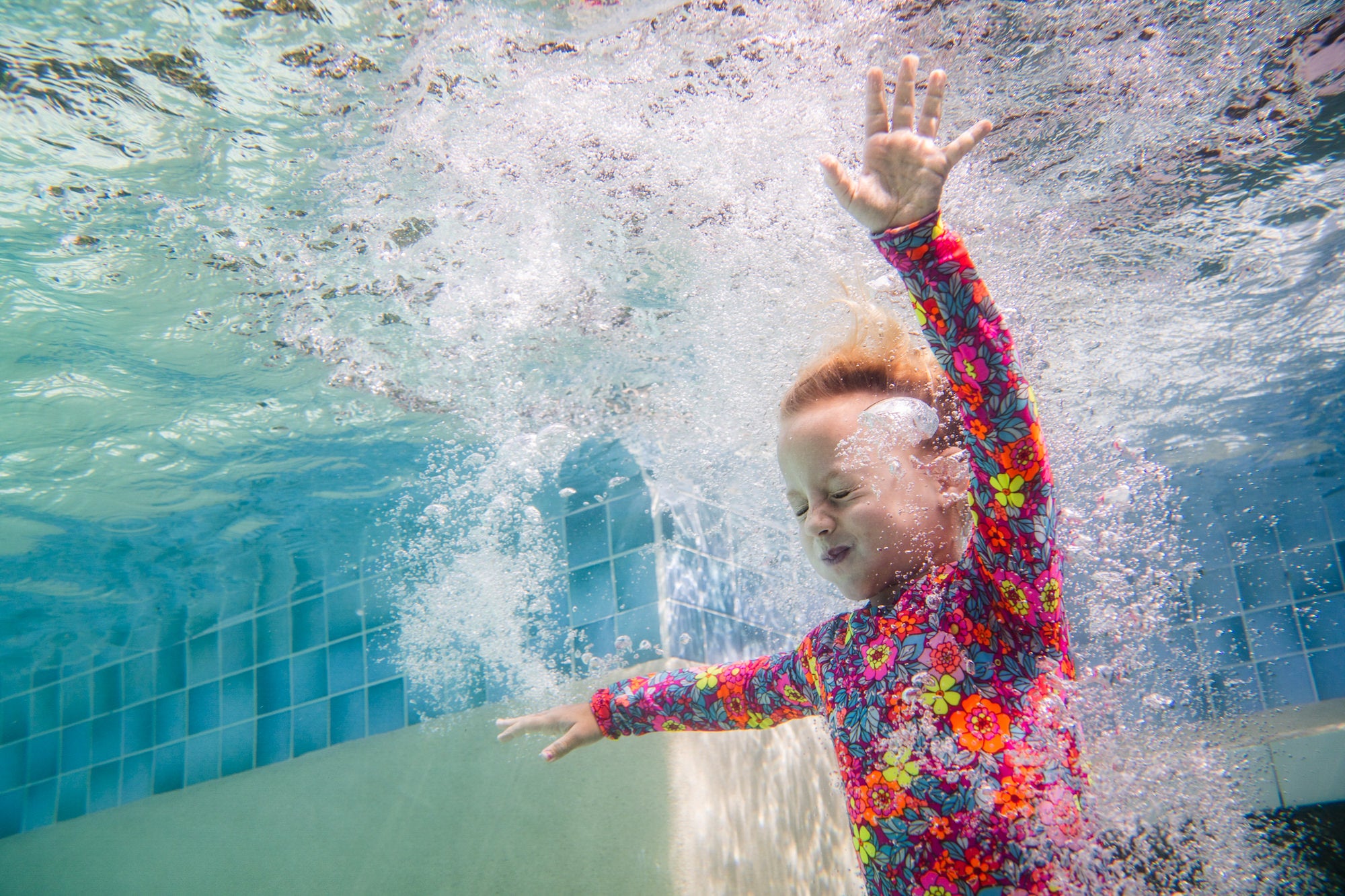 Girl splashes into the pool underwater, eyes closed, surrounded by bubbles near blue tiles.