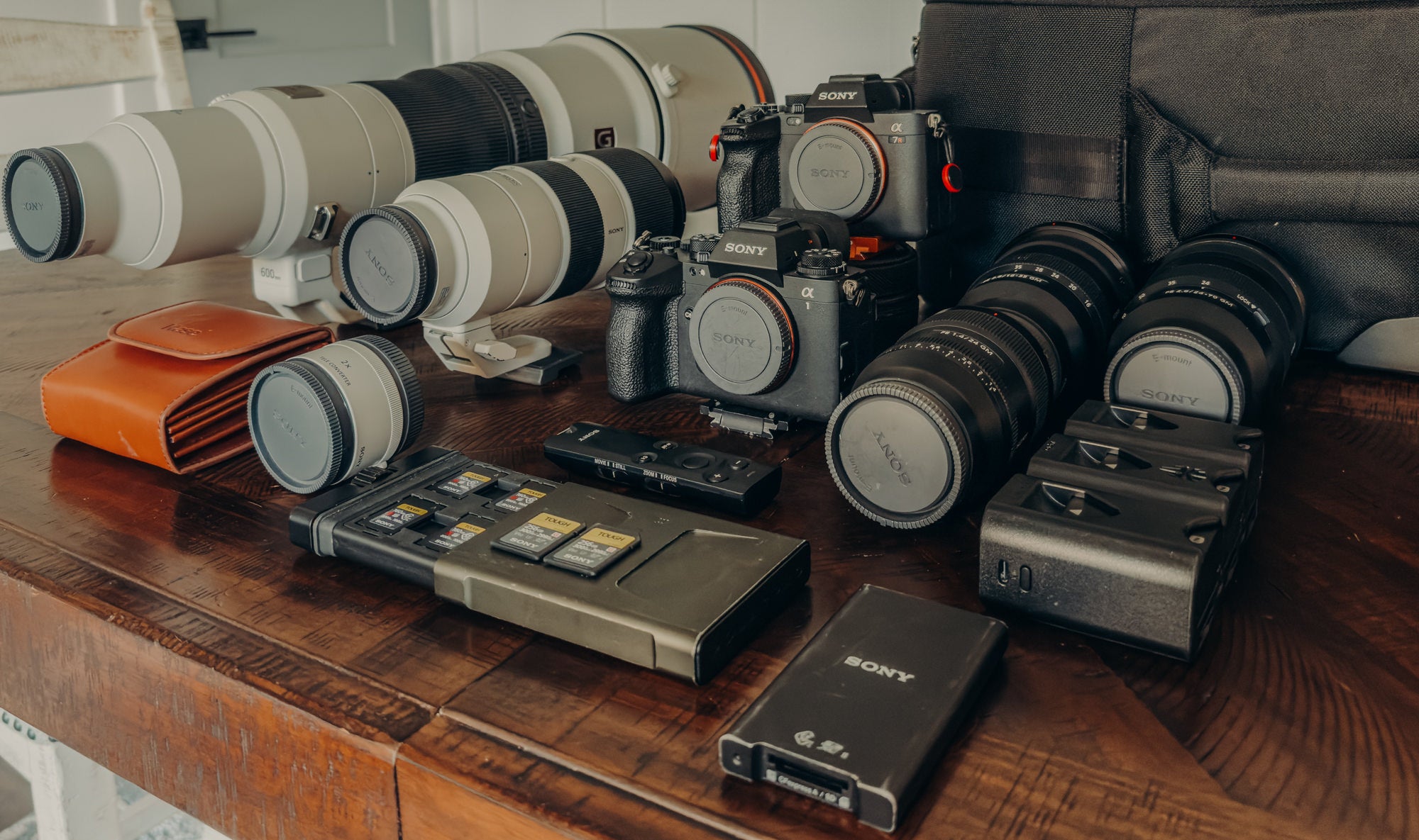 Sony Alpha camera bodies, white telephoto and black zoom lenses, memory cards and SSD laid out on a wooden table beside a camera bag.