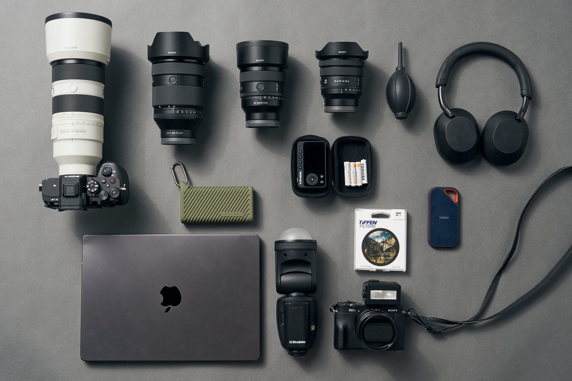 Flat lay of camera gear on gray: Sony Alpha body with telephoto lens, three G Master zooms, compact camera, flash, laptop, SSD, filter and headphones. Flat lay of camera gear on gray: Sony Alpha body with telephoto lens, three G Master zooms, compact camera, flash, laptop, SSD, filter and headphones.