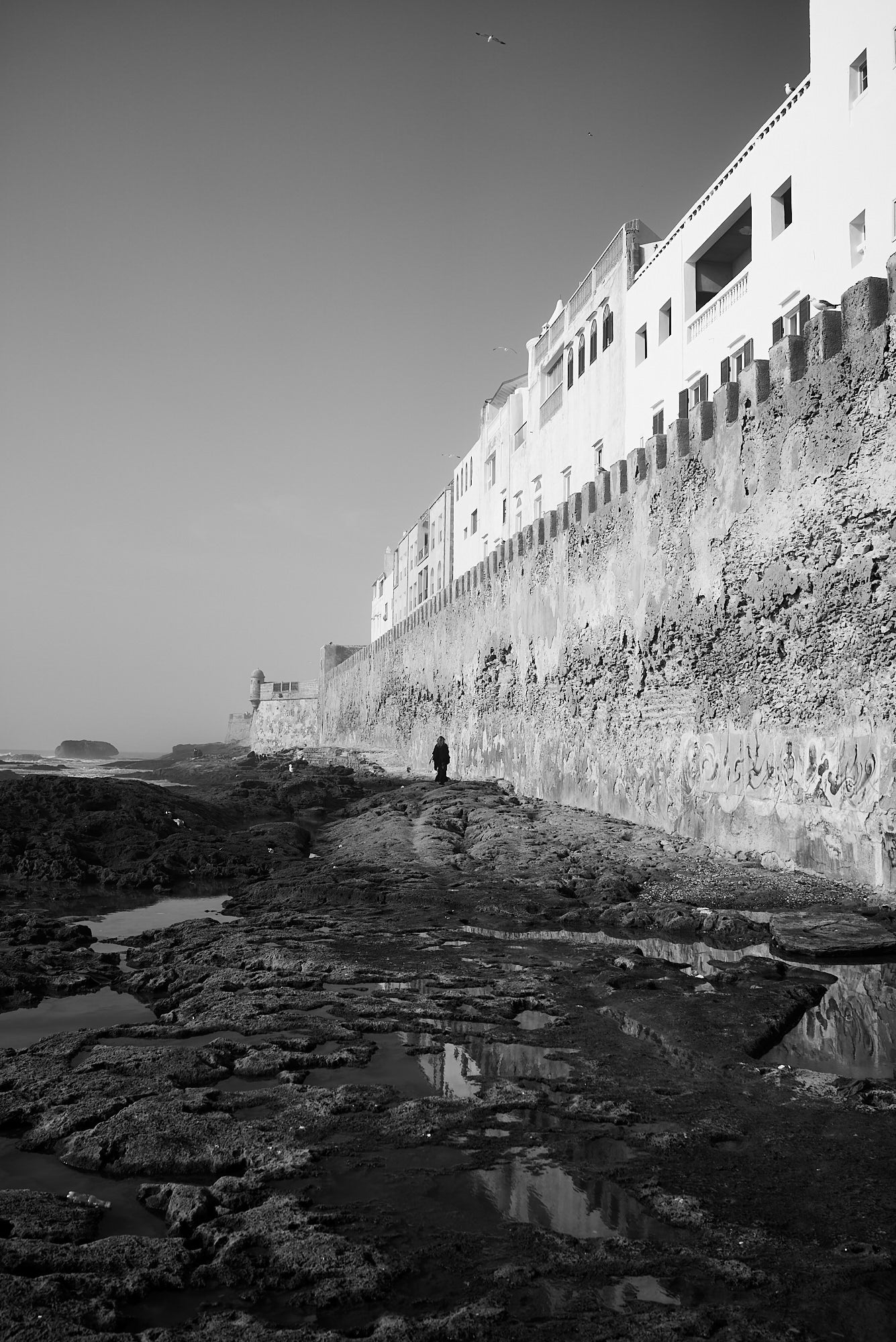 Black-and-white view of a lone figure walking rocky tidal flats beside a whitewashed seaside fortress wall. Photo by Nicholas Mastoras. RX1R III. 1/2500-sec. f/4.5. ISO 125. Black-and-white view of a lone figure walking rocky tidal flats beside a whitewashed seaside fortress wall. Photo by Nicholas Mastoras. RX1R III. 1/2500-sec. f/4.5. ISO 125.