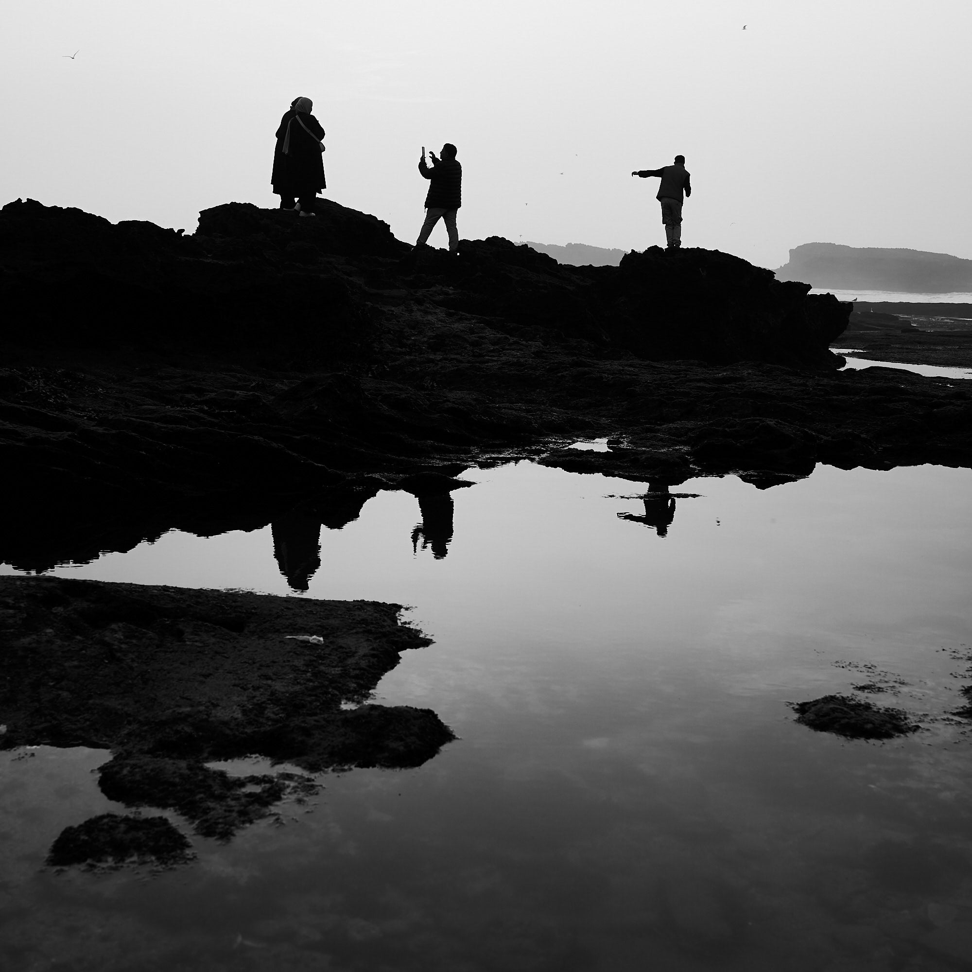 Three silhouetted figures on dark coastal rocks, mirrored in a still tide pool under a pale sky. Three silhouetted figures on dark coastal rocks, mirrored in a still tide pool under a pale sky.
