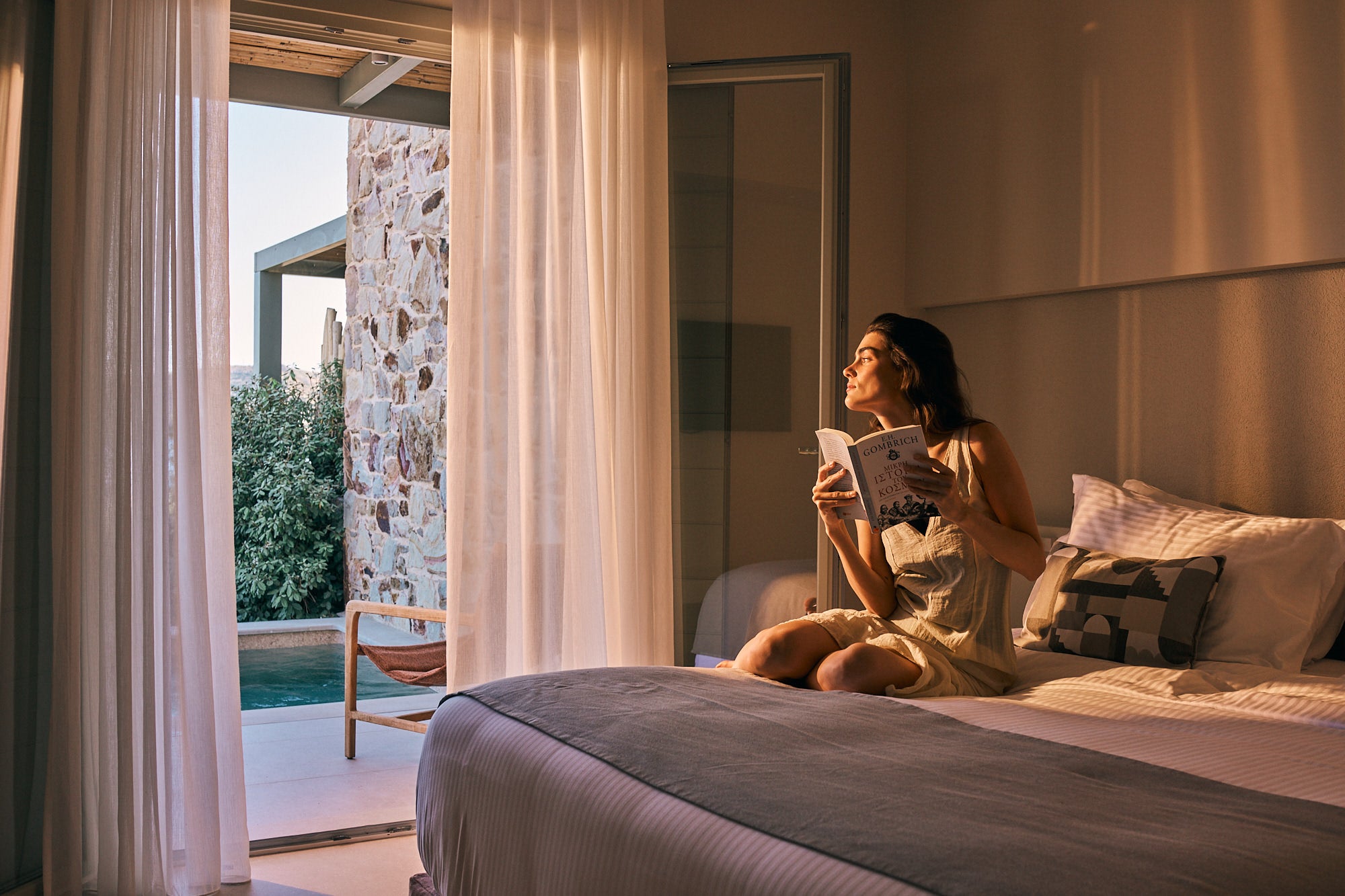 Woman sitting on a bed holding a book, gazing out an open doorway toward a stone wall and pool in warm light. Woman sitting on a bed holding a book, gazing out an open doorway toward a stone wall and pool in warm light.