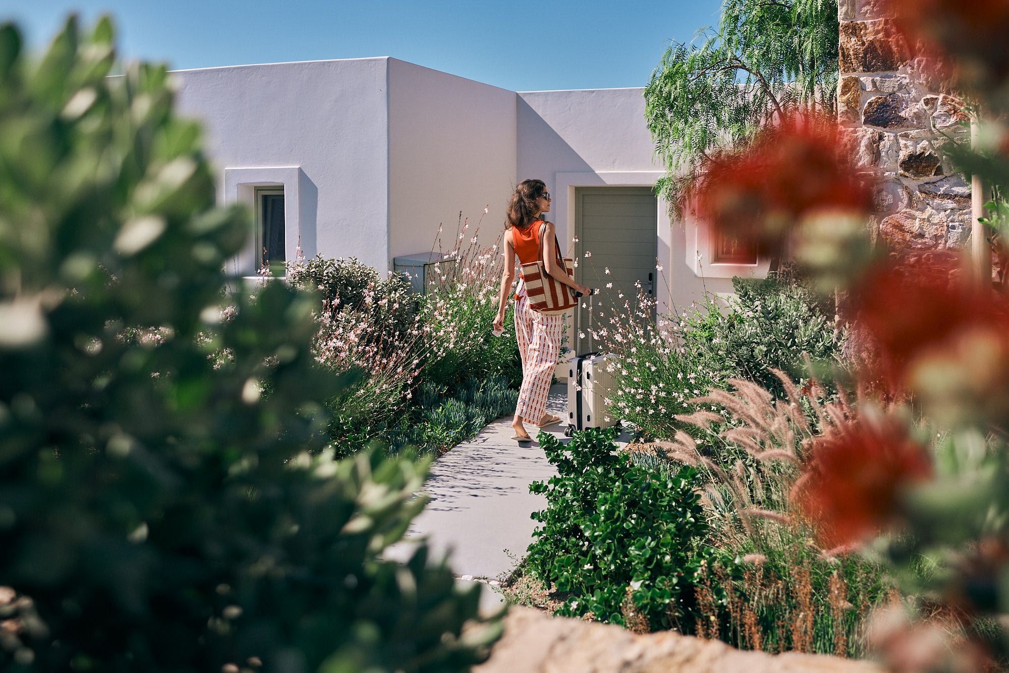 Woman pulling a suitcase along a garden path toward a whitewashed villa, framed by blurred flowering plants. Woman pulling a suitcase along a garden path toward a whitewashed villa, framed by blurred flowering plants.